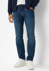 Dunkelblaue Denim-Jeans mit geradem Schnitt, ausgestattet mit fünf Taschen und subtilen Kontrastnähten, kombiniert mit weißen Sneakern.