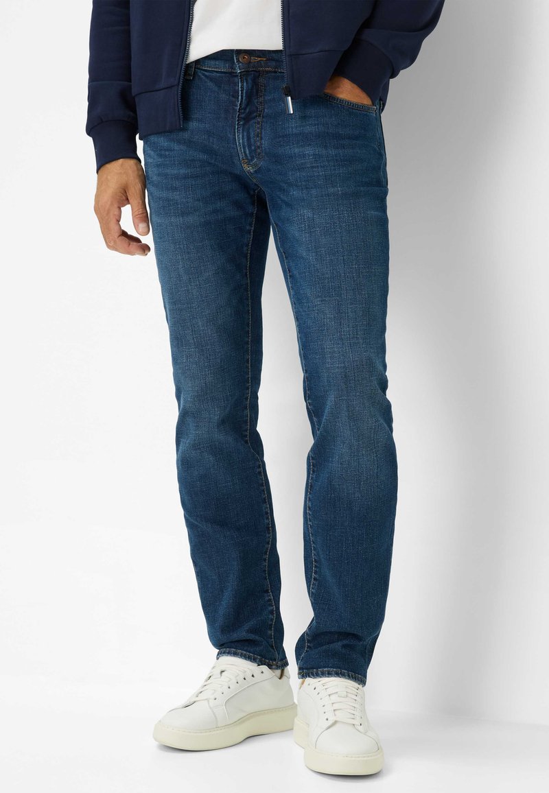 Dunkelblaue Denim-Jeans mit geradem Schnitt, ausgestattet mit fünf Taschen und subtilen Kontrastnähten, kombiniert mit weißen Sneakern.