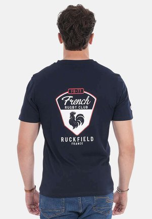 Homme portant un T-shirt bleu marine avec le logo "French Rugby Club" et un motif de coq dans le dos, associé à un jean bleu.