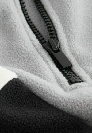 Grau Fleece-Stoff mit einem strukturierten Schwarz-Weiß-Muster. Besitzt einen schwarzen Reißverschluss, der ein elegantes, modernes Design unterstreicht.