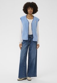 Frau mit lockigem Haar, die eine hellblaue Fleeceweste, ein weißes Langarmshirt, weit geschnittene blaue Jeans und cremefarbene Schuhe trägt. Sie steht vor einem weißen Hintergrund.