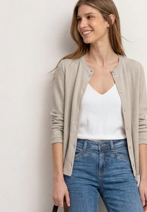 Smilende kvinde med lige, lyst brunt hår bærer en beige cardigan, hvid V-hals top og blå jeans, mens hun holder i en brun taskes håndtag ved sin side.