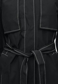 Μαύρο παλτό με ζώνη, λευκό ραφές αντίθεσης, μεγάλες εμπρός τσέπες με παπιγιόν και κάθετες ραφές στο σώμα.