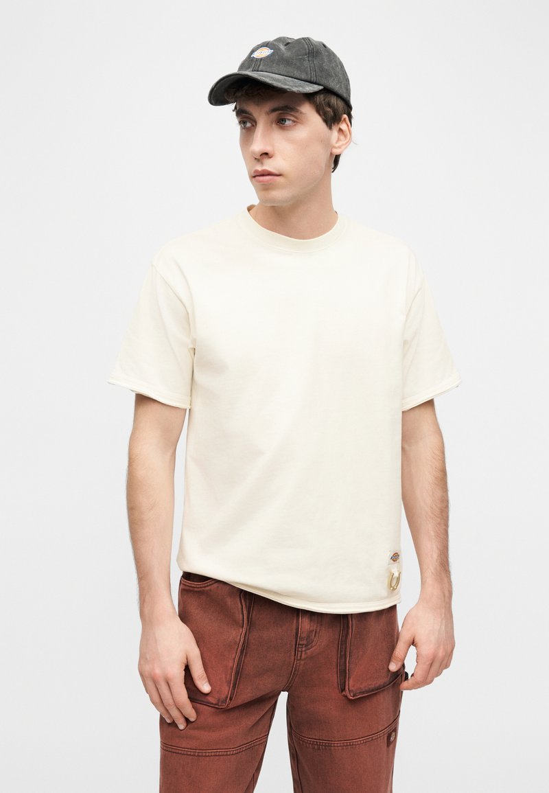 Junger Mann trägt ein cremefarbenes kurzärmeliges T-Shirt, rostfarbene Hosen mit großen Taschen und eine dunkelgraue Baseballkappe, stehend vor weißem Hintergrund.