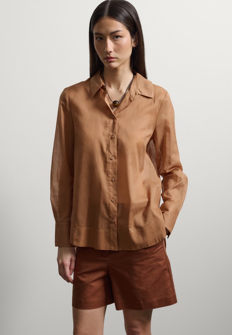 Mujer con una camisa de botones de manga larga de color marrón claro con cuello y pantalones cortos marrón oscuro, de pie con una mano en el bolsillo.