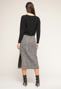 Schwarzer Strickpullover mit strukturiertem Muster und Knöpfen am Rücken, kombiniert mit einem knielangen Hahnentrittmuster-Rock mit einem Seitenschlitz. Schwarze Knöchelschäfte.