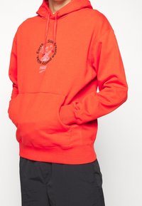 Röd hoodie tillverkad av bomullsblandning med framficka, justerbar huva med dragsko och ett grafiskt tryck med text på bröstet. Slimmad passform.