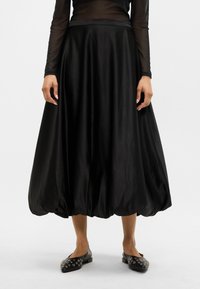 Fekete ballon szoknya sima anyagból, illeszkedő derékkal és dús, levegős szegéllyel. Fekete lapos cipőkkel párosítva, melyeket tűk díszítenek.