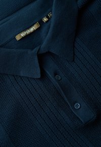 Sötétkék pólóing texturált kötésből, bordás gallérral, két gombos záródással és "SUPERDRY" feliratú textilmintával.