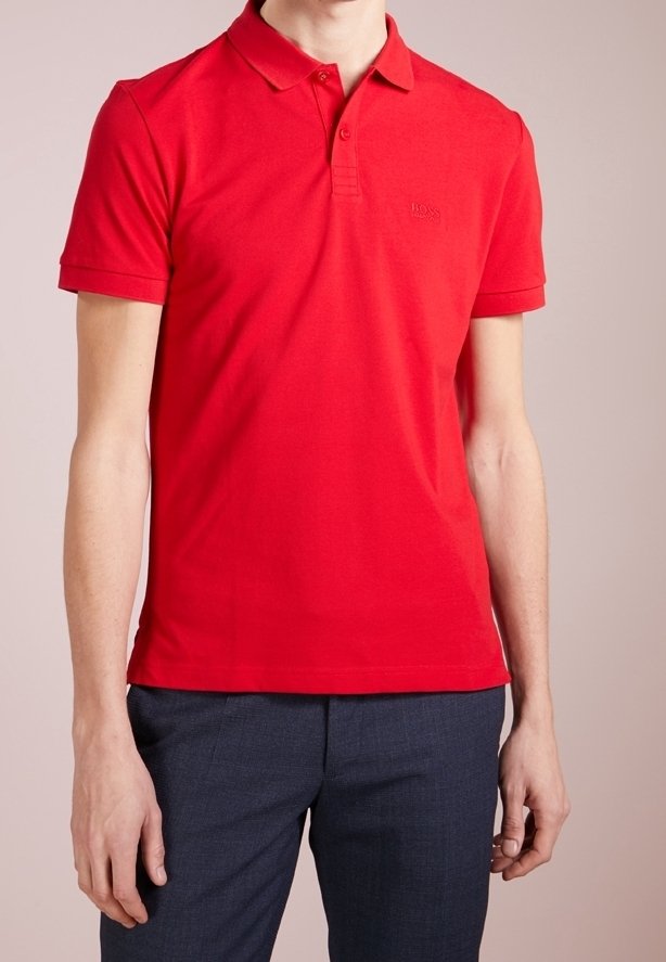 Homme portant un polo rouge uni à manches courtes avec col et un pantalon gris foncé, debout devant un fond neutre.