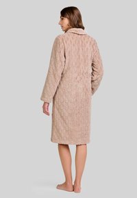 Mujer de pie, descalza, de lado, vistiendo un albornoz beige con textura hasta la rodilla, de mangas largas y con cuello.