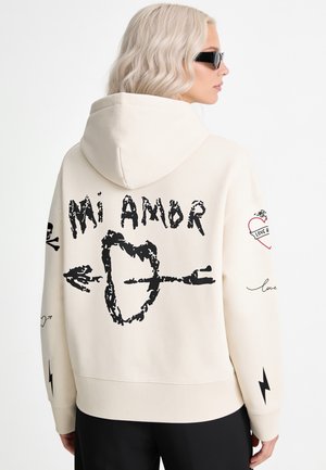 Frau mit kurzen blonden welligen Haaren und Sonnenbrille, die einen cremefarbenen Hoodie mit schwarzer Grafik "Mi Amor" und einem Pfeil durch ein Herz auf dem Rücken trägt.