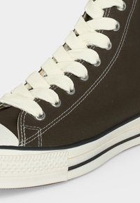Κοντινό πλάνο ενός σκούρου καφέ ψηλού καμβά sneaker με λευκά κορδόνια, διπλή λευκή ραφή και λευκή λαστιχένια σόλα με μαύρη ρίγα.
