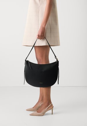 Borsa hobo in pelle nera con finitura testurizzata, forma curva, manico corto e dettaglio del logo in oro. Tenuta in mano con scarpe con tacco beige.