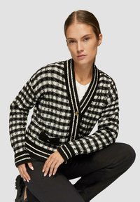 Schwarz-weiß kariertes Strickjacke mit V-Ausschnitt, goldenen Knöpfen und kontrastierenden gestreiften Kanten. Das Material wirkt strukturiert und gemütlich.