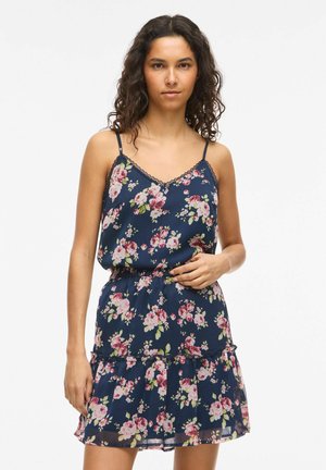 Femme aux cheveux bouclés portant une robe sans manches bleu marine à fleurs avec de fines bretelles, debout devant un fond blanc uni.