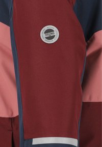 Utomhusjacka med en kombination av vinröda, marinblå och rosa tygpaneler. Har en rund metallisk logotyp och en reflexrem.