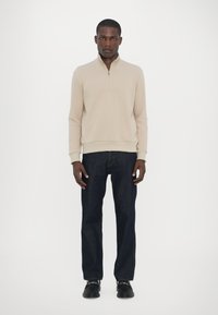 Beige zip-up sweatshirts med hög krage, ihop med mörkblå raka jeans och svarta skor. Enkel design, avslappnad passform.