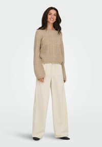 Pull beige tricoté avec motif texturé, manches larges, associé à un pantalon large crème et des chaussures plates noires.