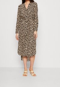 Robe à manches longues à imprimé léopard, avec ceinture nouée à la taille, col en V et fente latérale. Confectionnée en tissu léger, portée avec des sandales de couleur claire.