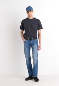 Camiseta gris, jeans azules, cinturón negro y zapatos negros. Modelo lleva una gorra azul, de pie contra un fondo blanco.