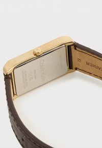 Χρυσή ορθογώνια κάσα ρολογιού με καφέ υφασμάτινο λουράκι. Διαθέτει κορώνα και εγχάρακτο λογότυπο στην πίσω πλευρά από ανοξείδωτο χάλυβα.