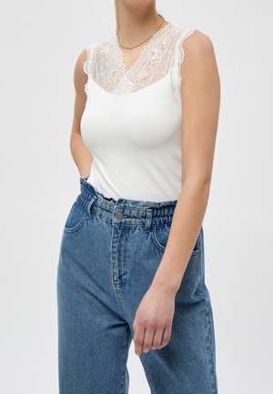 Femme portant un haut blanc sans manches avec un décolleté en dentelle et un jean bleu taille haute, debout devant un fond clair uni.