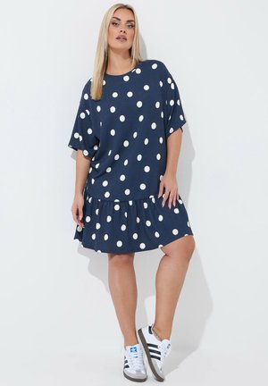 Kobieta stojąca, ubrana w granatową sukienkę w białe kropki oraz białe buty sportowe Adidas z czarnymi paskami.