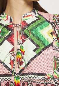 Multikleurige jurk met een patroon van geometrische vormen in groen, rood en roze, met een kraag en versierde kralenstrikjes.