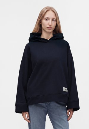 Femme aux longs cheveux blonds portant un sweat à capuche noir ample et un jean bleu, debout devant un fond clair uni.
