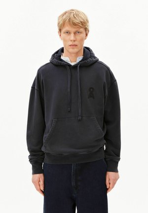 Blonder Mann trägt einen schwarzen Kapuzenpullover mit Fronttasche und dunklen Jeans, steht vor einem weißen Hintergrund.