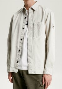 Vaaleanharmaa napillinen paita, joka on valmistettu puuvillasta, rintatasku, mustat napit ja pieni logopatchi hihassa.