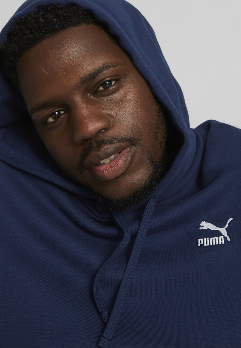 Marineblå hættetrøje lavet af blødt stof, med en kengurulomme, justerbar snor og hvidt Puma-logo på venstre brystområde.