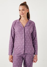 Violet pyjamapuku valkoisilla pilkuilla. Pitkähihainen paita, jossa on kaulus ja nappikiinnitys; pehmeä, tekstuuriinen kangas. Housujen yksityiskohdat vastaavat.