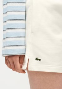 Χέρι που ακουμπά δίπλα σε λευκή φούστα με πράσινο λογότυπο κροκόδειλου· ορατό πουκάμισο με ριγέ μοτίβο σε ανοιχτό μπλε, λευκό και γκρι με μακριά μανίκια.