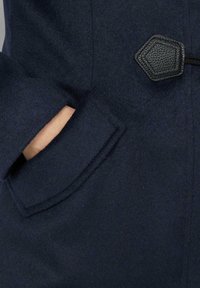 Hand teilweise in der Tasche eines marineblauen Wollmantels mit Leder-Details am oberen rechten Rand.