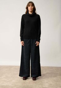 Schwarzer Pullover mit Kapuze, kombiniert mit weit geschnittenen, strukturierten dunklen Hosen. Das Outfit zeichnet sich durch eine lockere Passform und eine glatte, weiche Textur aus. Braune Schuhe runden den Look ab.