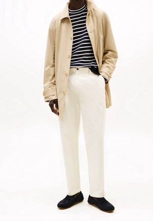 Imperméable beige, t-shirt rayé bleu marine et blanc, pantalon blanc et chaussures bleu marine. Dispose de boutons sur le manteau et d'une coupe structurée et ample.