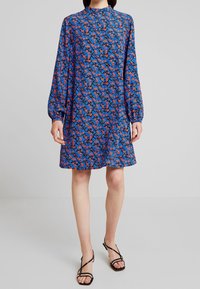 Robe à manches longues en tissu bleu avec un motif floral rouge et bleu. Elle présente un col montant et une coupe décontractée, se terminant au-dessus des genoux.