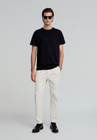 Tricou negru cu guler rotund, asortat cu pantaloni bej deschis și pantofi negri. Modelul poartă ochelari de soare închiși la culoare, stând în fața unui fundal neutru.