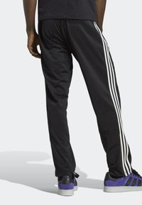 Pantalones deportivos negros con rayas blancas a los lados, con una textura suave y cinturilla elástica, combinados con zapatillas moradas.