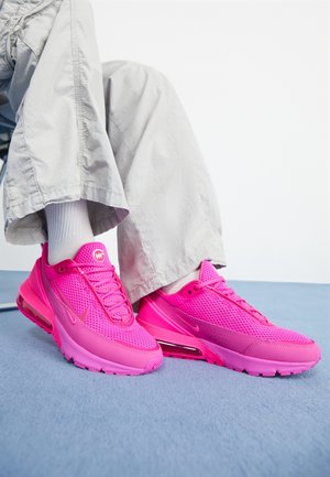 Osoba ubrana w jasnobeżowe spodnie i białe skarpetki, siedząca, prezentująca jaskrawo różowe buty Nike na niebieskiej podłodze na białym tle.