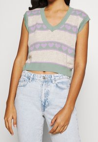 Gebreide mouwloze trui met een V-hals, voorzien van een lichtbeige basis, groene en lavendel hartpatronen en geribbelde accenten. Casual cropped pasvorm.