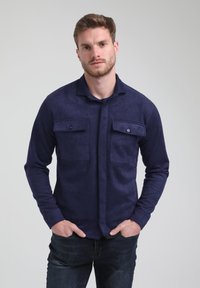 Camicia a maniche lunghe di un blu navy, realizzata in un materiale simile al suede, con due tasche sul petto, chiusura con bottoni e taglio rilassato, indossata sopra jeans scuri.