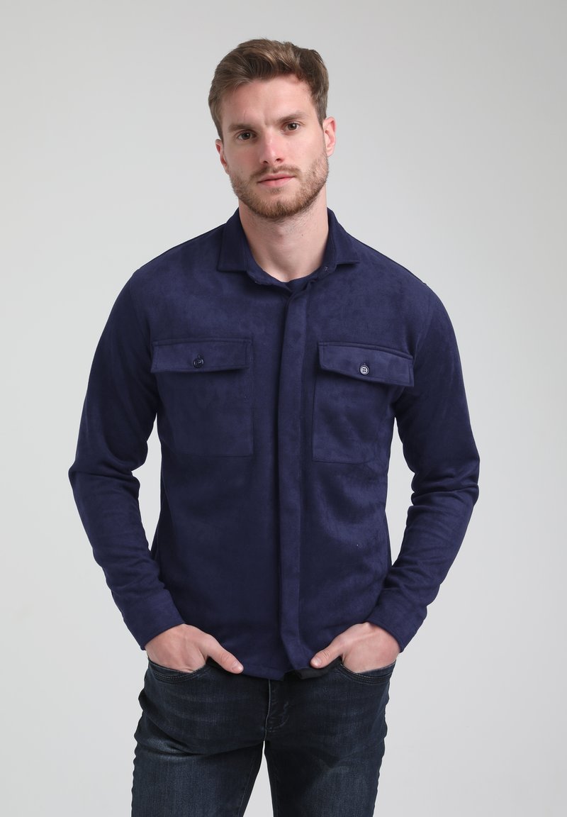 Camicia a maniche lunghe di un blu navy, realizzata in un materiale simile al suede, con due tasche sul petto, chiusura con bottoni e taglio rilassato, indossata sopra jeans scuri.