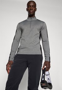 Sivý pánský svetr s dlouhým rukávem a částečným zipem, texturovaná látka a černé boční panely. Model má na sobě tmavé sportovní kalhoty a drží popruh.