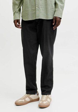 Personne portant un pantalon noir, des baskets beige et blanches, et une chemise vert clair boutonnée, debout devant un fond uni.