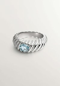 Anillo de banda retorcida de plata con una gema ovalada de color azul claro. El diseño presenta texturas suaves y redondeadas, y un acabado pulido.