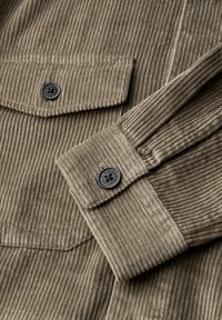 Olivově zelená kordová látka s vertikálním žebrováním, s dvěma náprsními kapsami a černými knoflíky, která se vyznačuje strukturovaným povrchem.