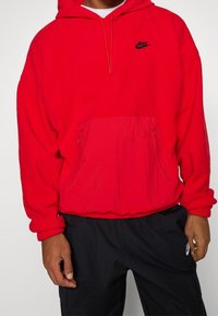 Felpa rossa realizzata in tessuto morbido con una tasca frontale tipo kangaroo e due tasche laterali con zip. Presenta un logo Nike nero sul petto.
